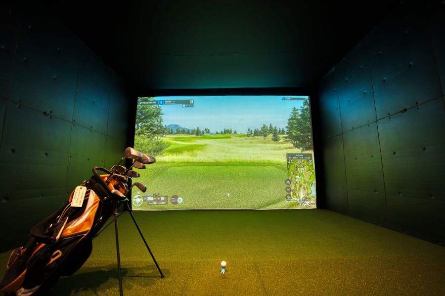 Home golf simulator setup featuring a large projection screen displaying a golf course, with a golf bag and clubs positioned on green turf in front of the screen.